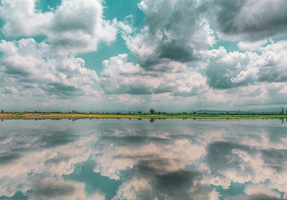 Ein wolkenverhangener Himmel, darunter ein Stück Land. Unterhalb ein See, in dem sich die Wolken spiegeln.